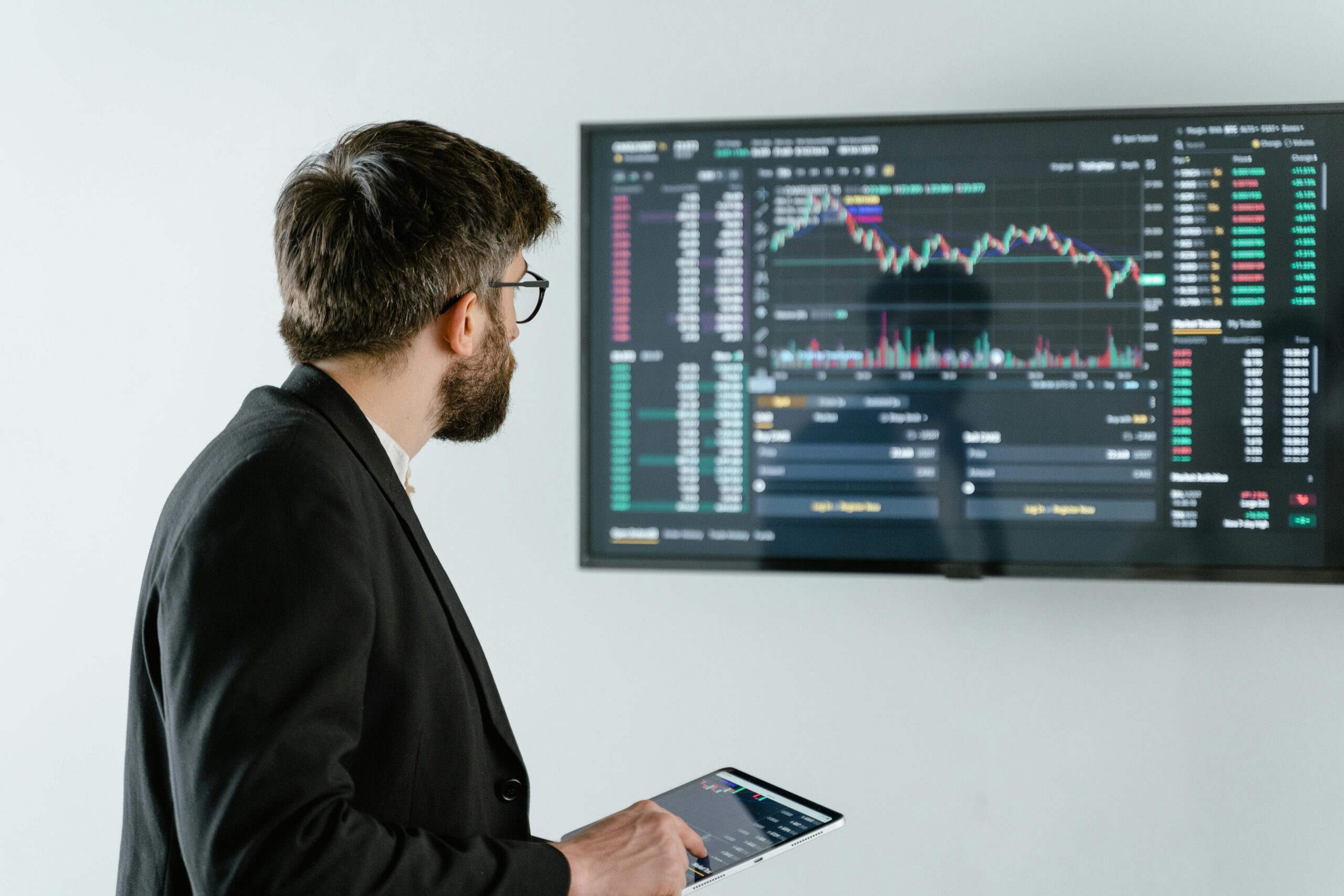 Man observing stock market data on a screen and holding a tablet, representing financial analysis and trading.